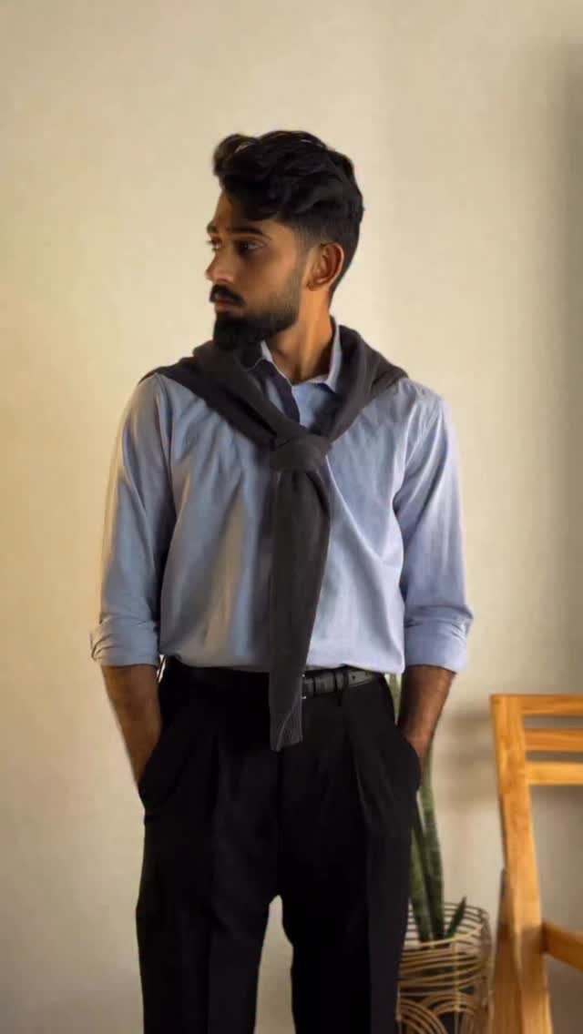 A man wearing a light blue shirt, black trousers, and a grey sweater tied around his shoulders.