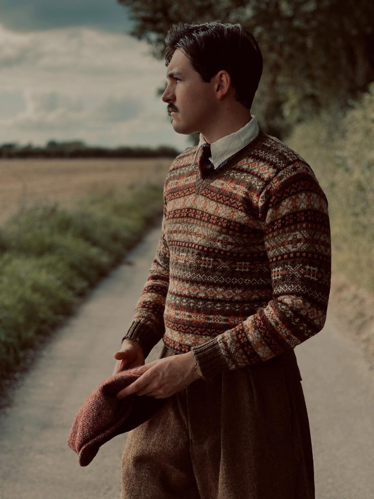 Man wearing a patterned Fair Isle sweater and collared shirt standing on a country path.