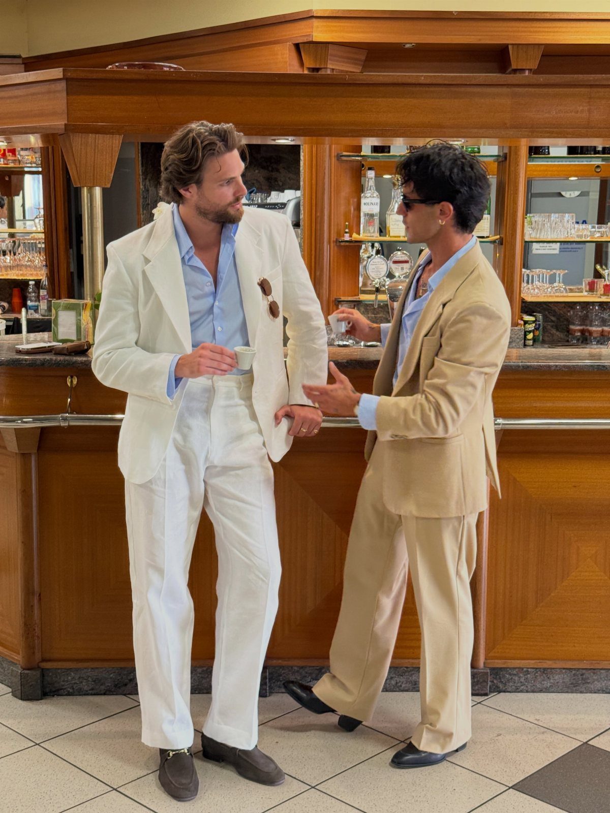 Two men in light-colored summer suits standing and talking by a wooden bar.