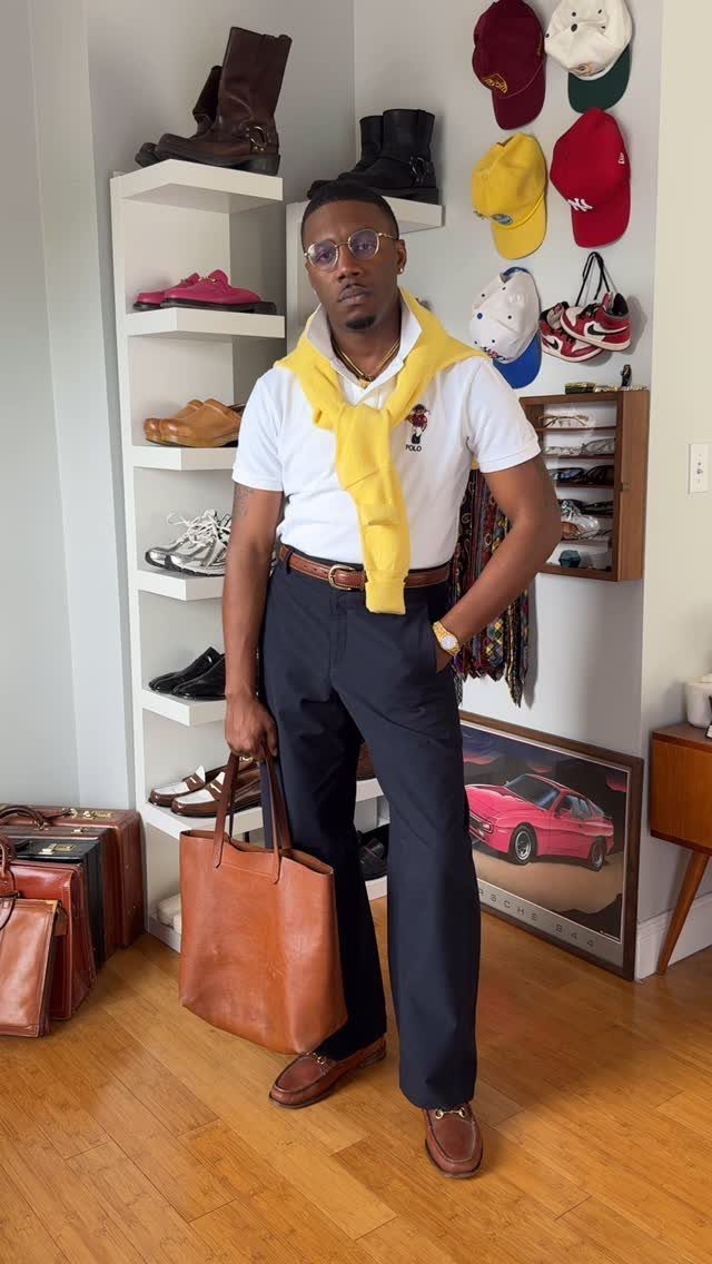 Man in a white polo shirt, yellow sweater, navy trousers, and brown loafers holding a leather tote.