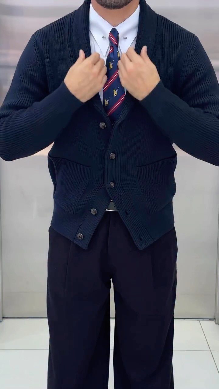 Man wearing a navy shawl cardigan, white shirt, patterned tie, and dark trousers.