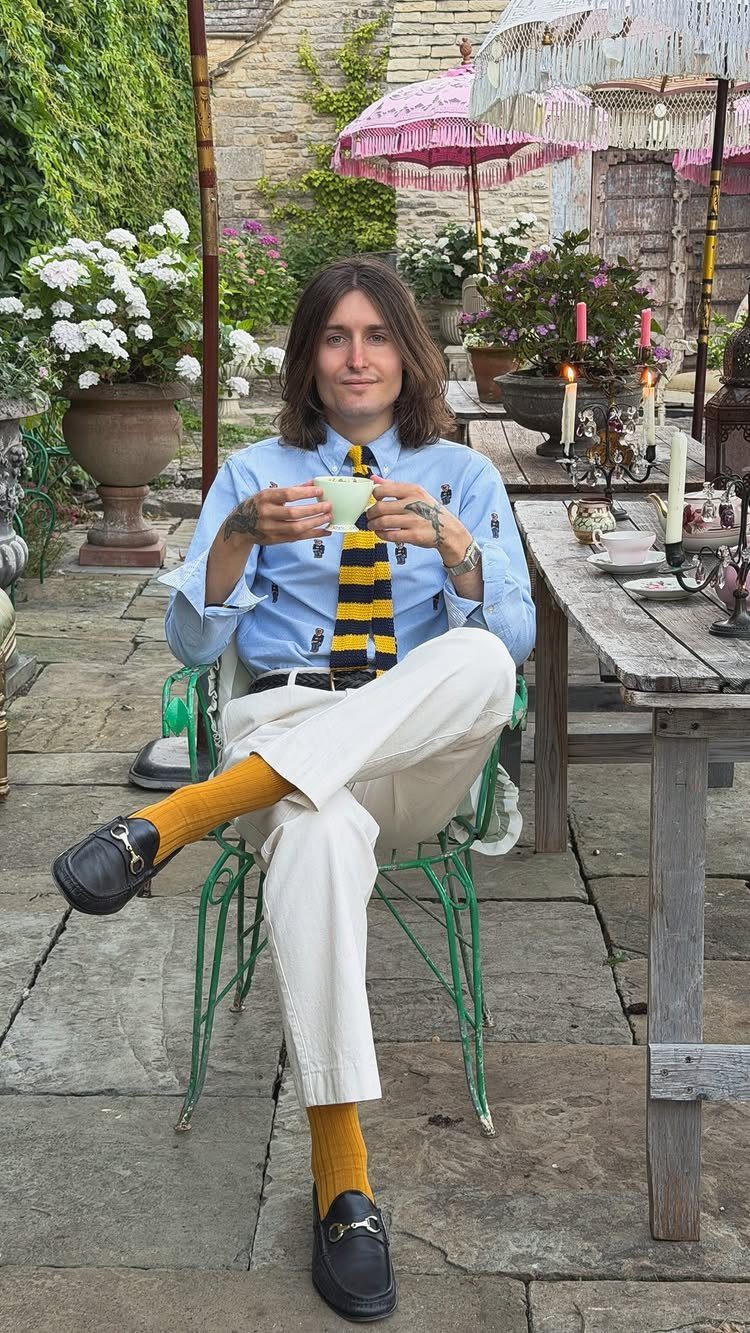 Man in preppy attire sitting in a garden holding a teacup.
