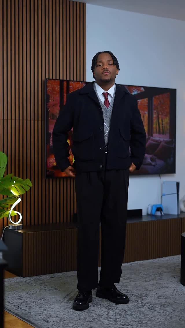 Man posing in a preppy outfit consisting of a navy jacket, sweater vest, tie, and black trousers.