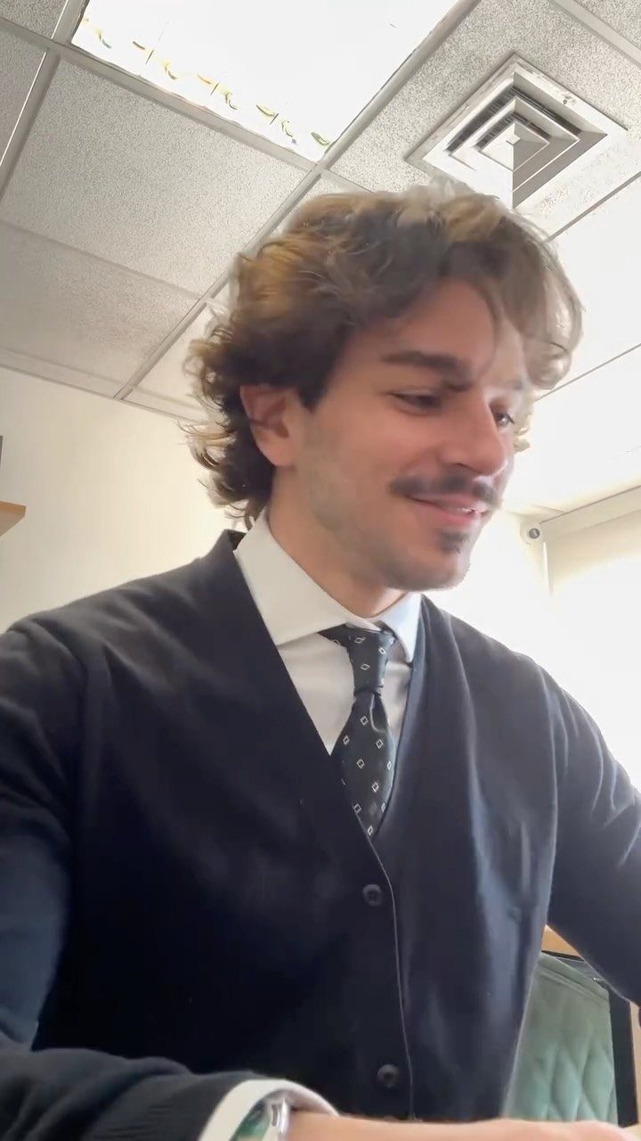 Man with wavy hair wearing a white shirt, patterned tie, and black cardigan in an office.