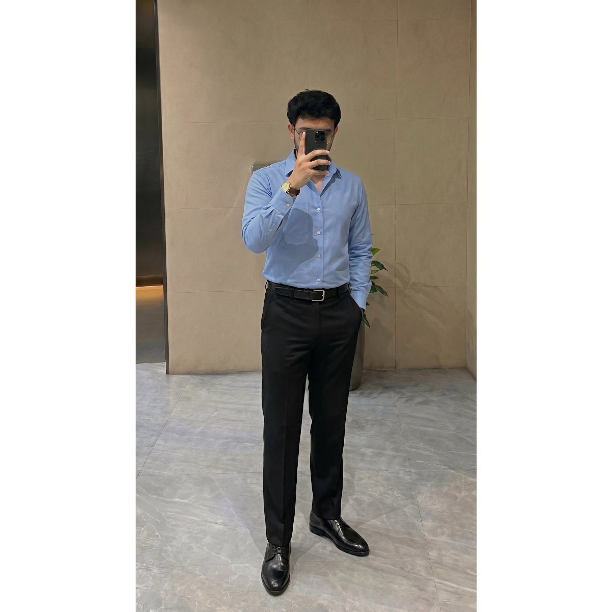 Man in a light blue button-down shirt and black trousers posing for a mirror selfie.