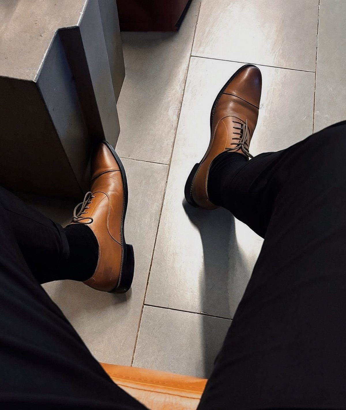Tan leather derby shoes worn with black trousers and socks on a tiled floor.