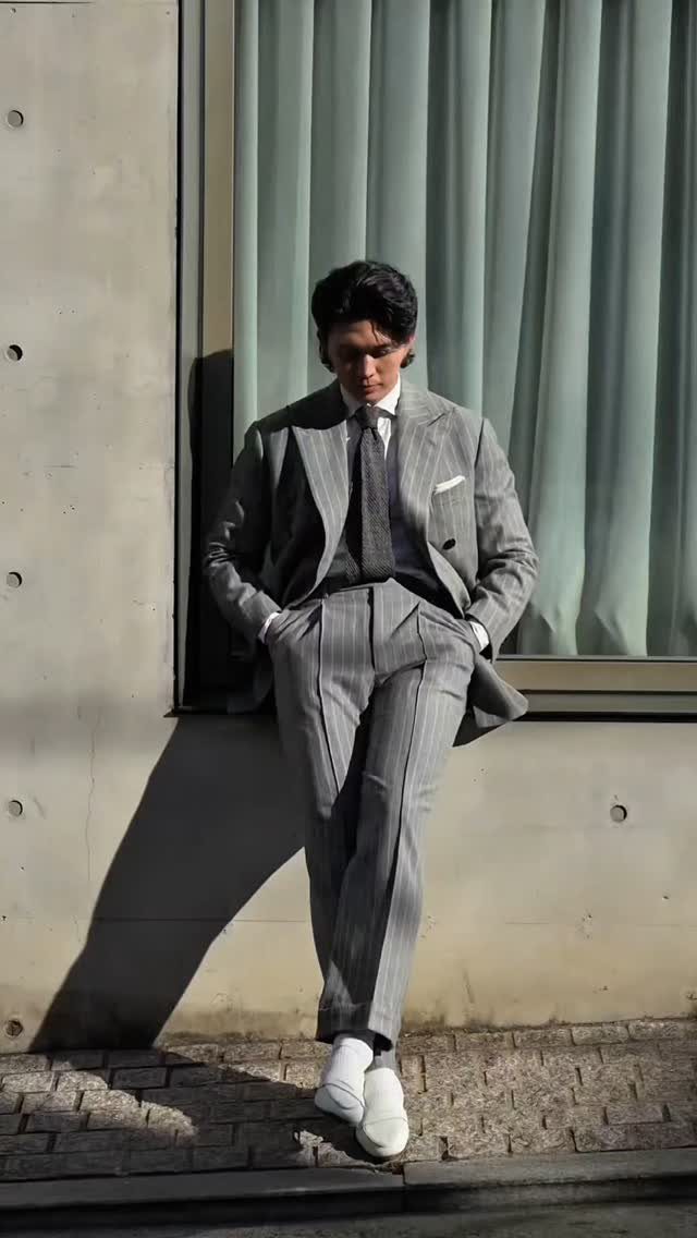 A man in a grey pinstripe tailored suit leaning against a wall outdoors.
