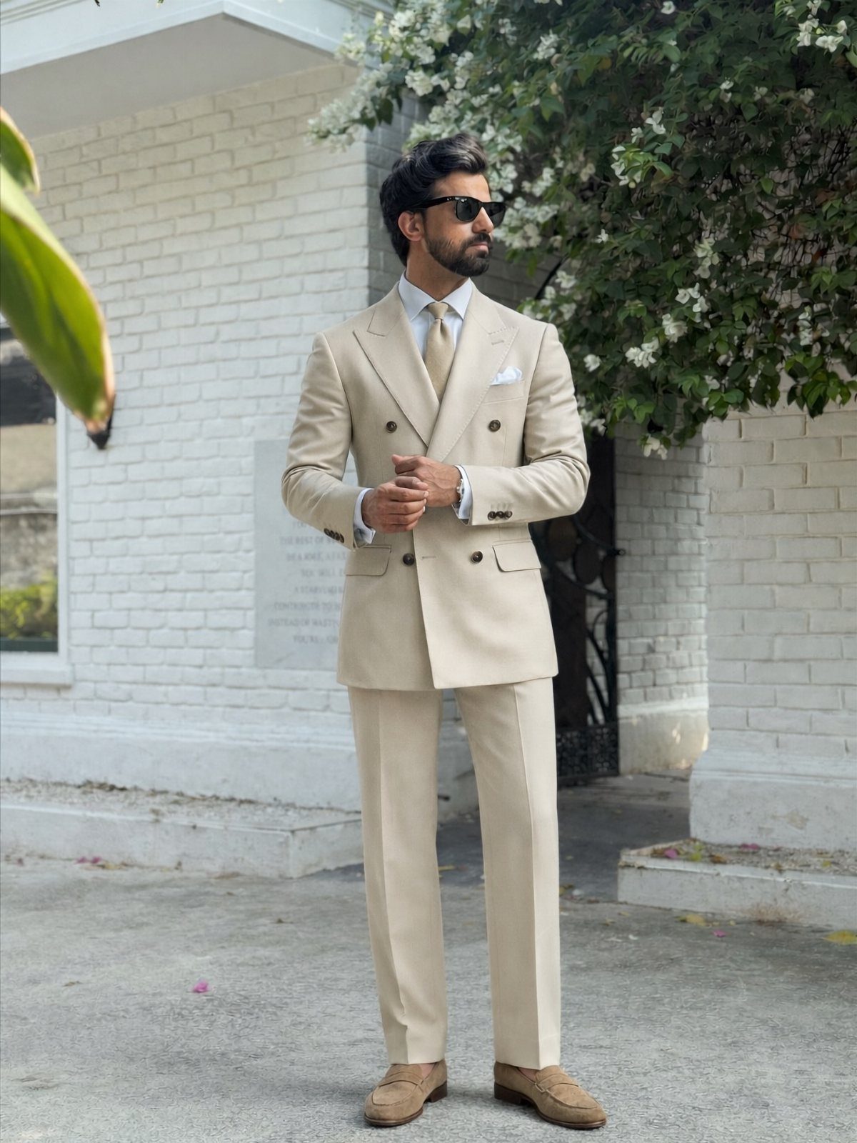 Man in a beige double-breasted suit and sunglasses standing by a white brick wall.