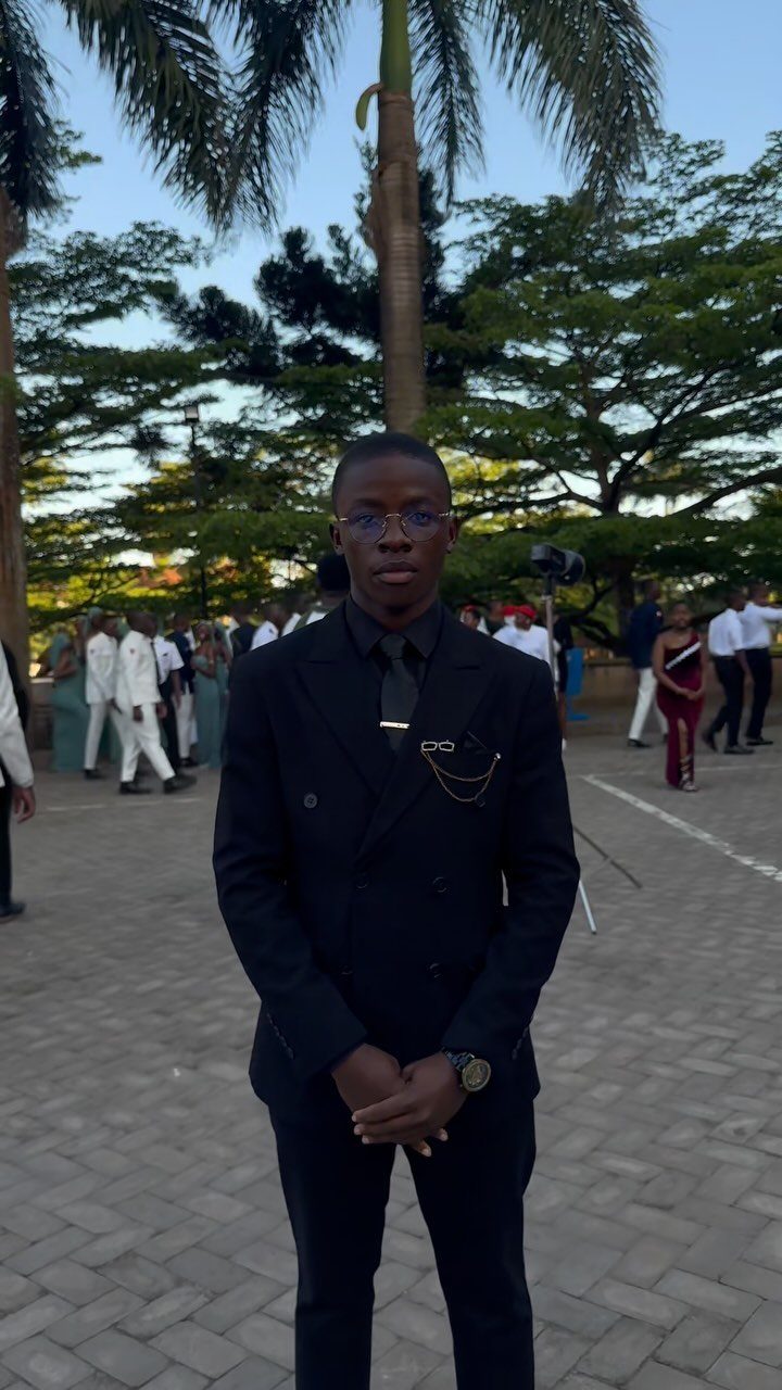 A young man posing in a black double-breasted suit and black shirt outdoors.