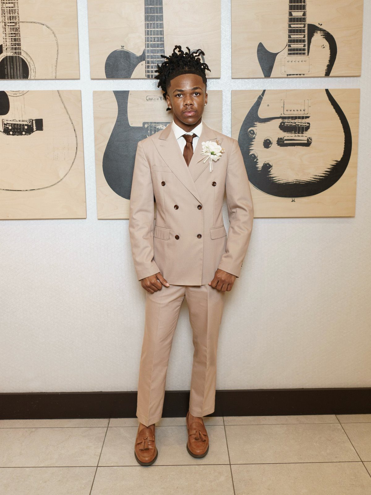 A young man posing in a tan double-breasted suit with a brown tie and white boutonniere.