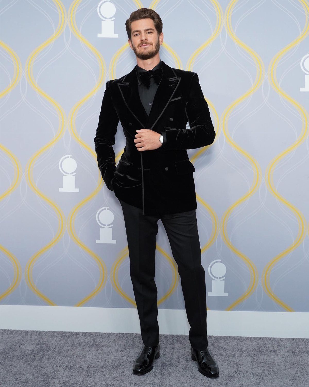 A man wearing a black velvet blazer, black bowtie, and black trousers standing against a patterned backdrop.