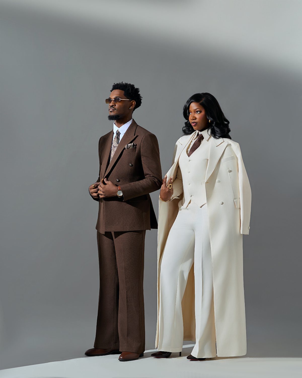 A man in a brown suit and a woman in a white suit standing against a grey background.