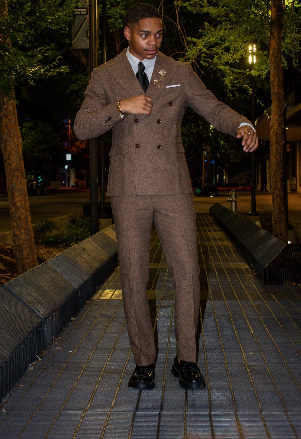 A young man posing in a brown textured double-breasted suit on a city walkway at night.