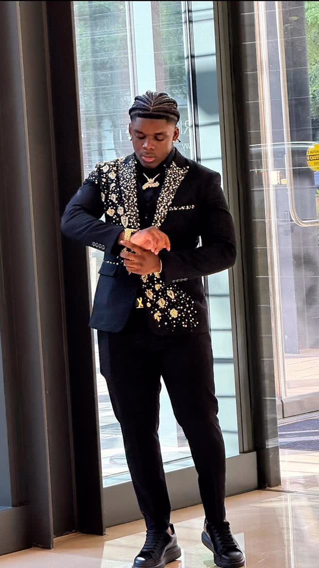 A young man posing in a black suit with ornate gold and silver embroidery on the lapels.