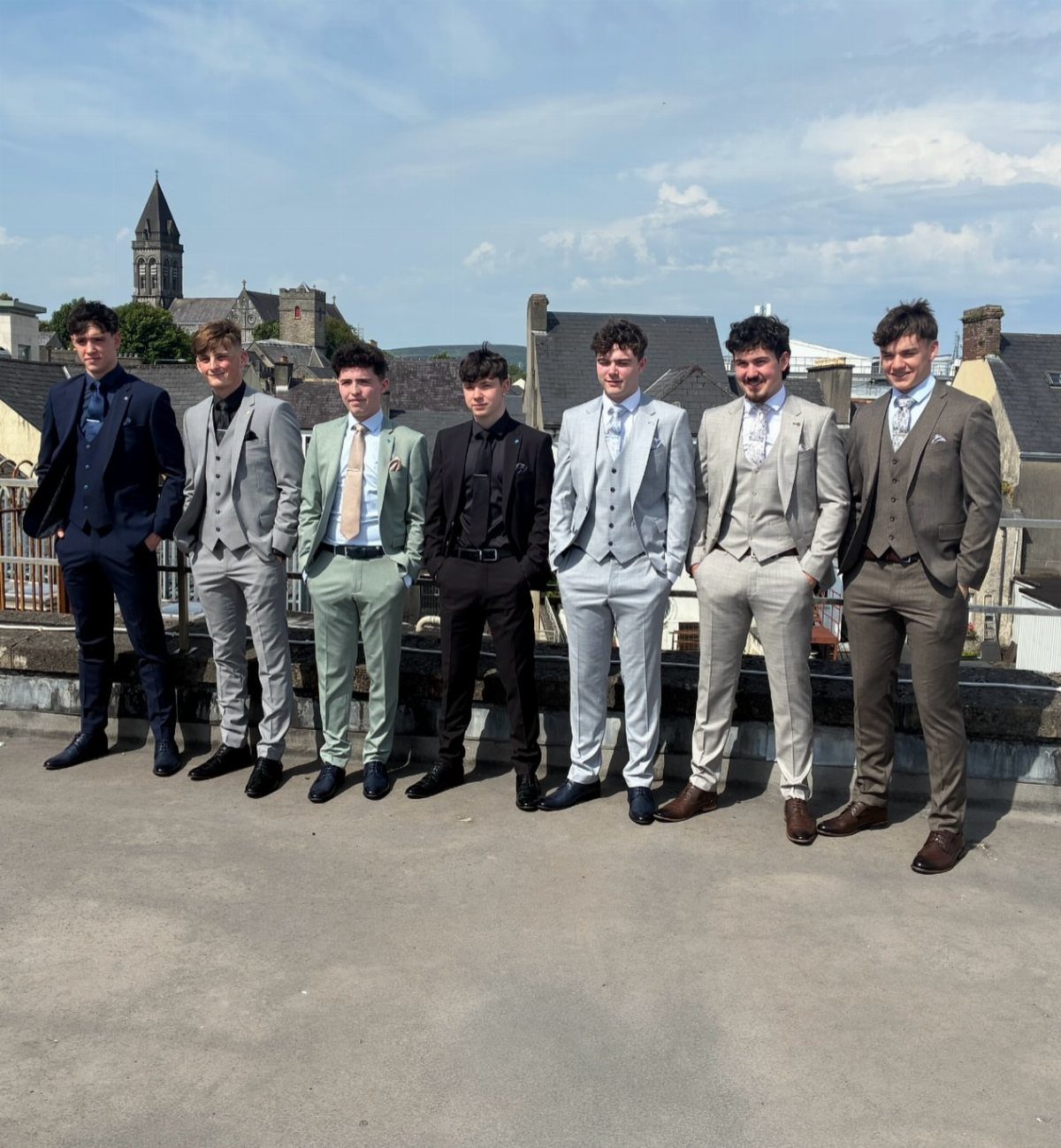 Seven young men posing outdoors in various colored three-piece suits on a rooftop.