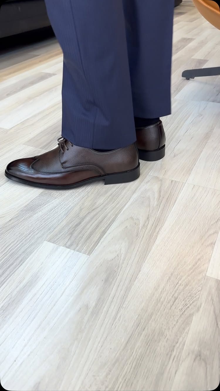 Brown leather derby shoes worn with navy blue trousers on a light wood floor.