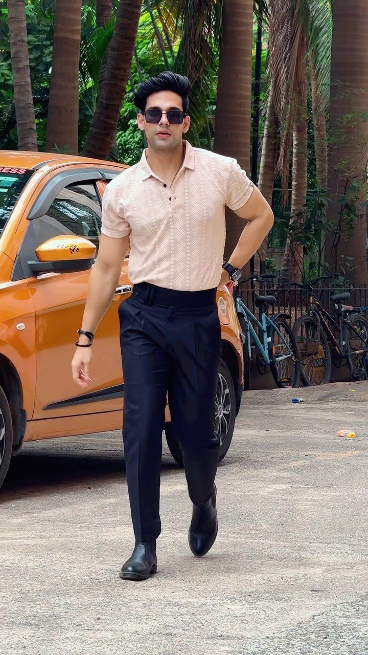 Man walking in a beige polo shirt and navy tailored trousers near an orange car.