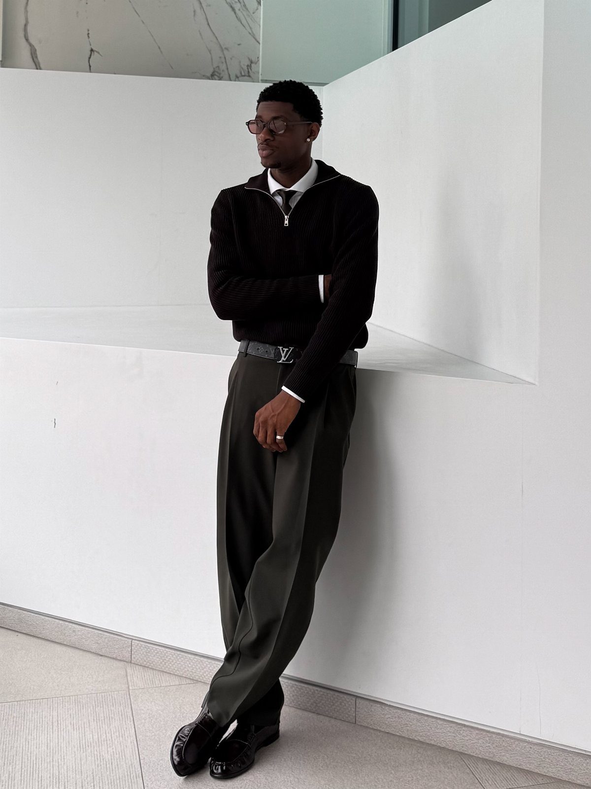 A man wearing a black sweater, tie, and olive trousers leaning against a white wall.