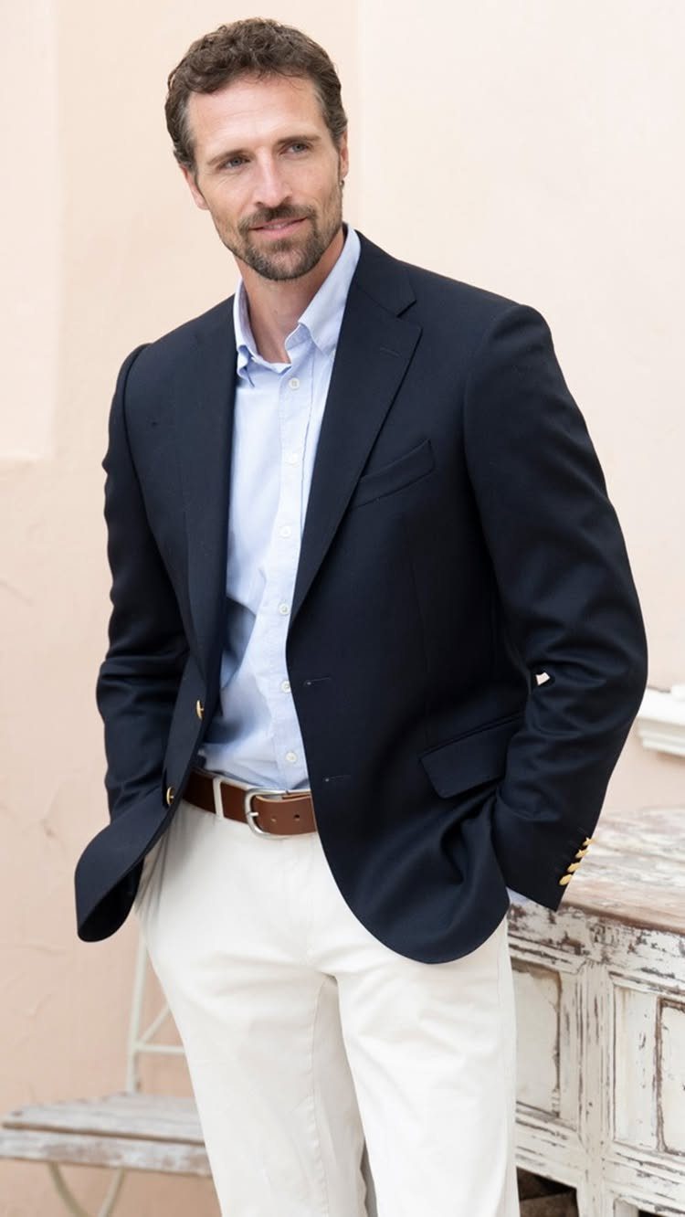 A man wearing a navy blazer, light blue shirt, and white trousers standing outdoors.