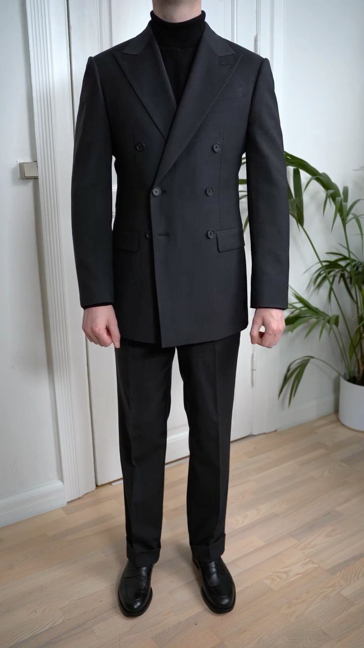 Man wearing a black double-breasted suit, black turtleneck, and black dress shoes standing indoors.