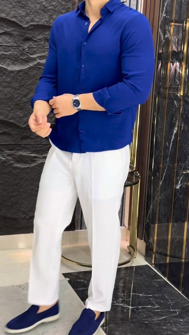 Man wearing a blue button-down shirt, white trousers, and navy slip-on shoes standing indoors.