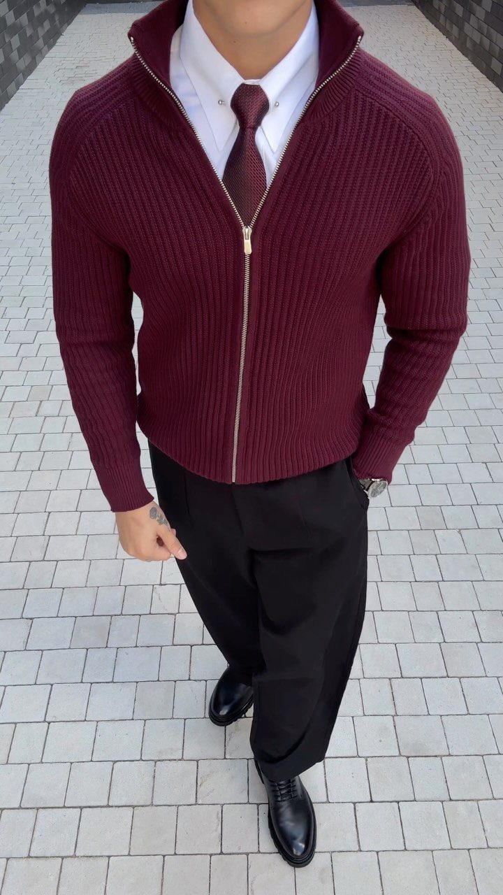 Man wearing a burgundy ribbed cardigan, white shirt, tie, and black trousers standing outdoors.