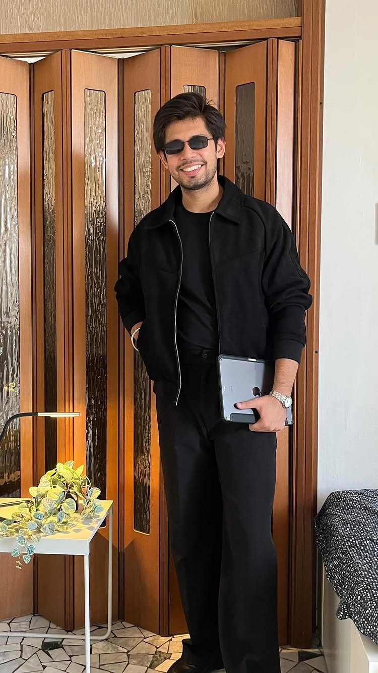 A man in a black jacket and sunglasses smiling while holding a tablet indoors.