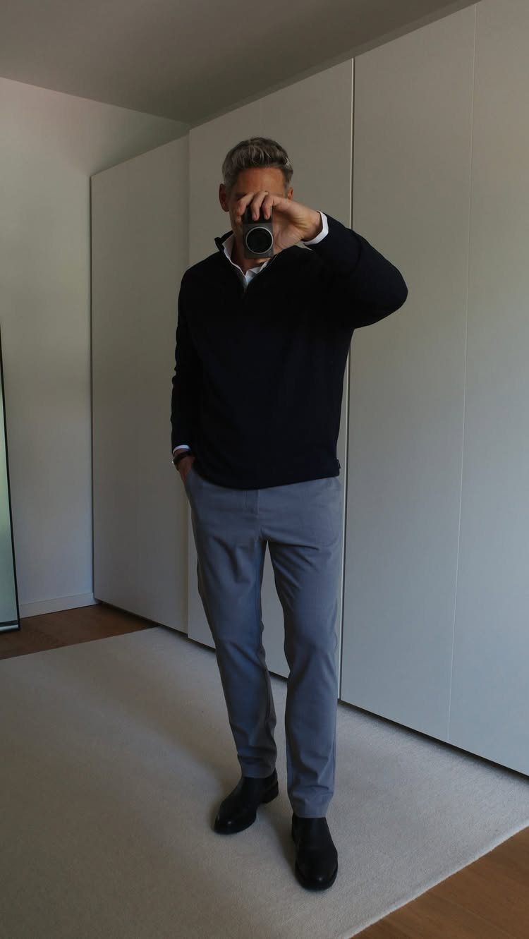 Man in navy pullover, light blue shirt, grey trousers, and black shoes taking a mirror selfie.