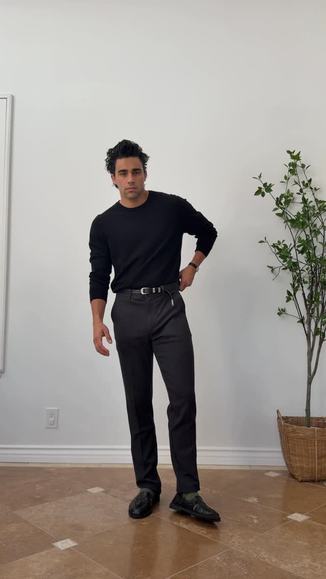 A man posing in a black sweater, dark grey trousers, and black loafers against a white wall.