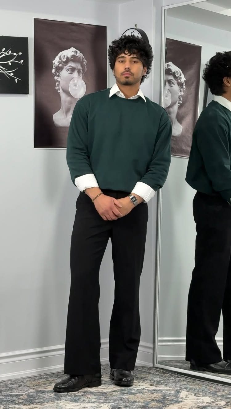 A man wearing a dark green sweater, white collared shirt, and black trousers standing indoors.