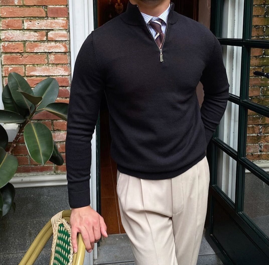 Man wearing a black quarter-zip sweater, white shirt, and beige trousers standing by a door.