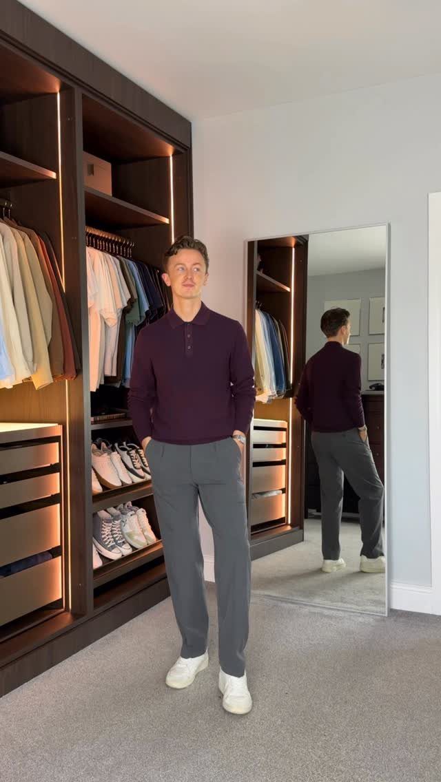 Man wearing a purple polo shirt and grey tailored trousers standing in a bedroom.
