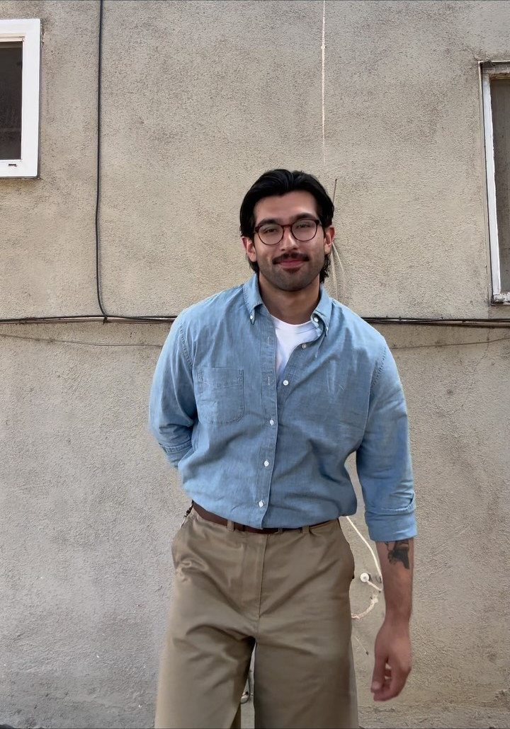 Man wearing a light blue chambray shirt and khaki chinos standing against a grey wall.