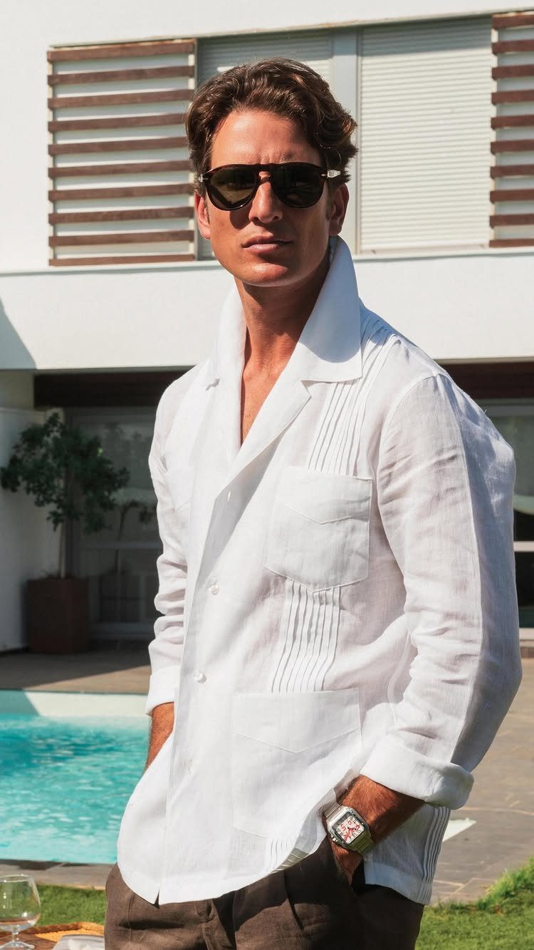 Man in sunglasses wearing a white linen Guayabera shirt standing by a pool.