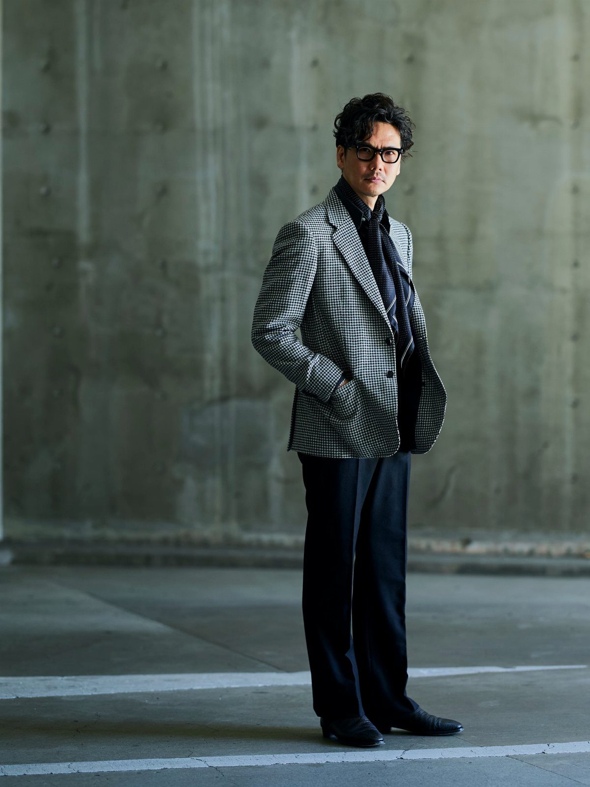 A man in a patterned sport coat, dark scarf, and black trousers standing against a concrete wall.