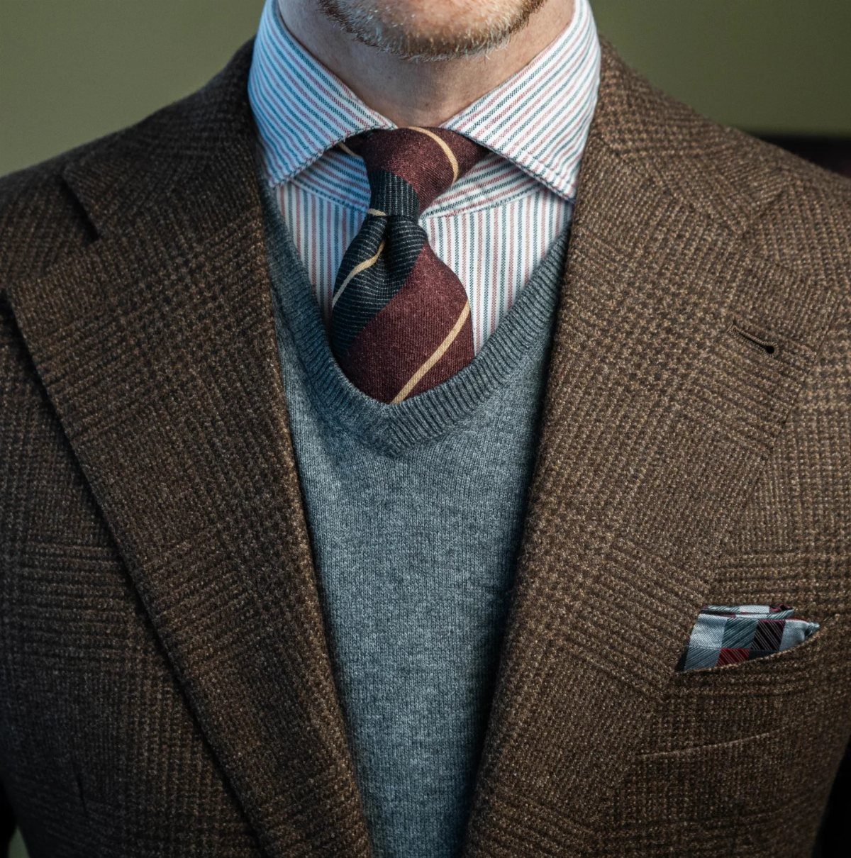 A man wearing a brown textured sport coat over a grey v-neck sweater and striped tie.