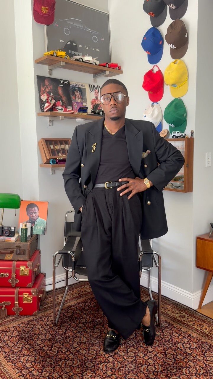 A man wearing an all-black monochromatic sport coat outfit and black trousers standing in a room.