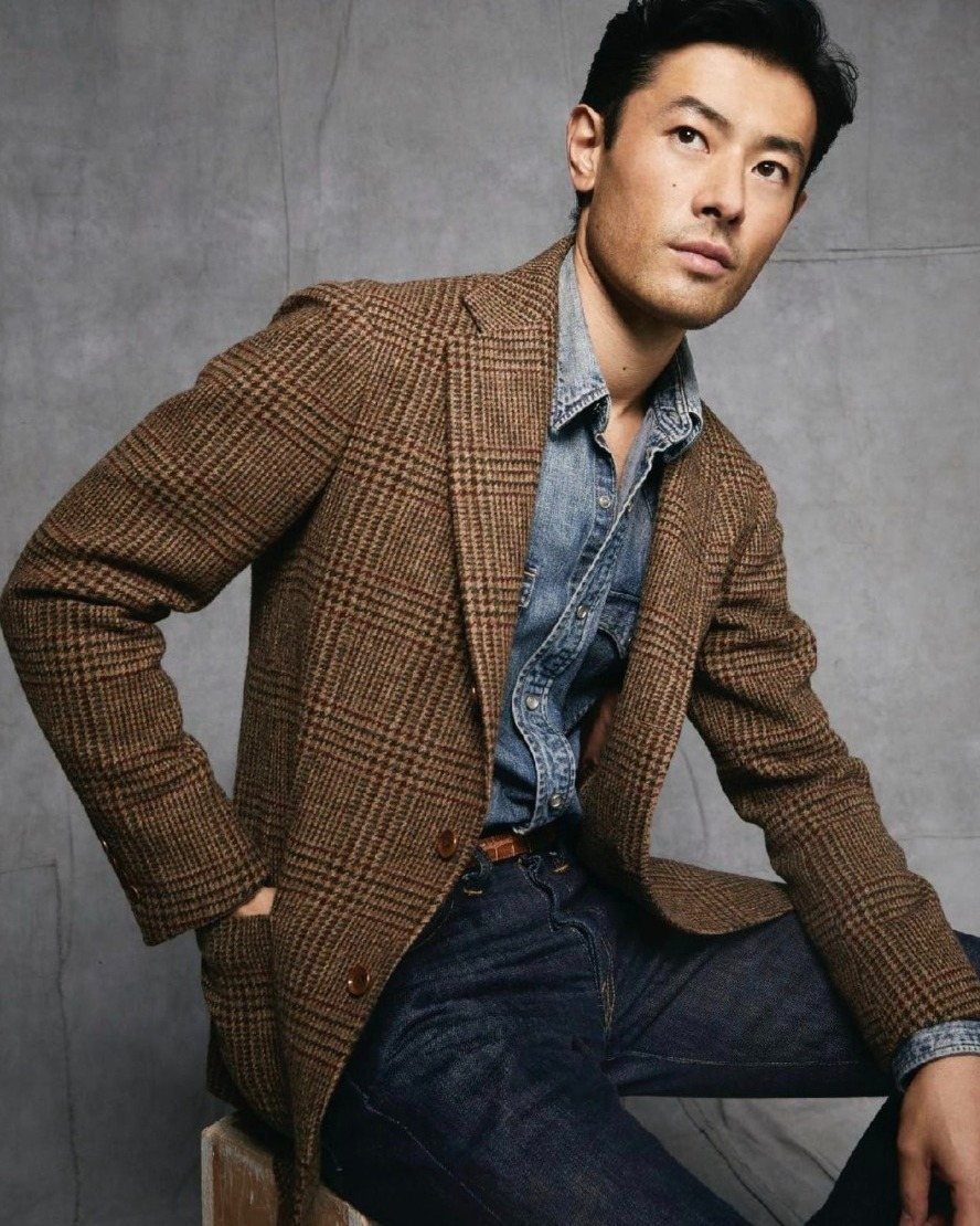 A man wearing a brown patterned sport coat, denim shirt, and dark blue jeans.