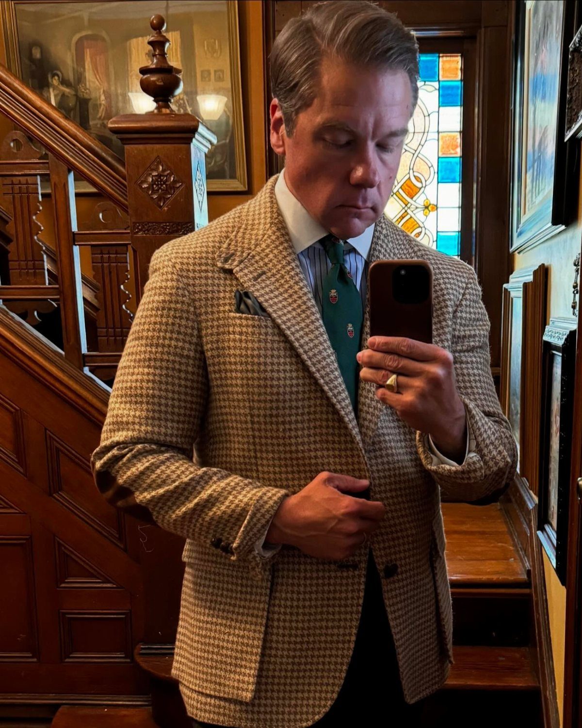 Man in a houndstooth sport coat and striped shirt taking a mirror selfie indoors.