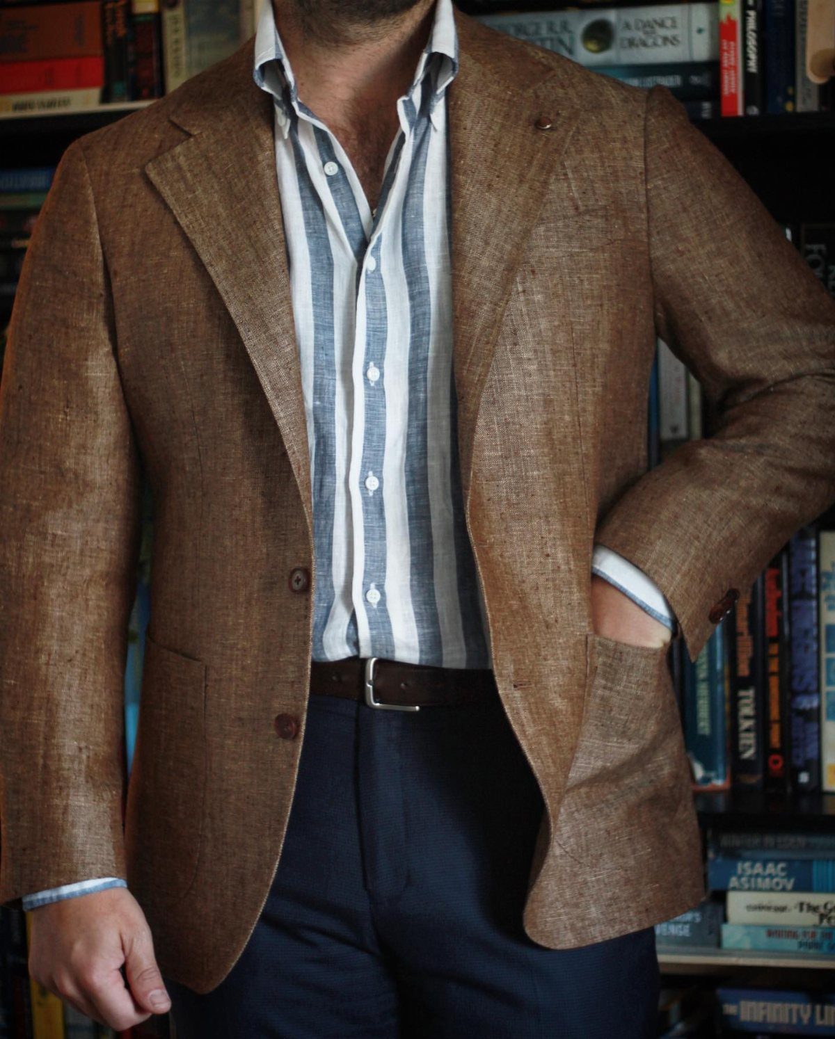 Man wearing a brown textured sport coat, striped shirt, and navy trousers standing before bookshelves.