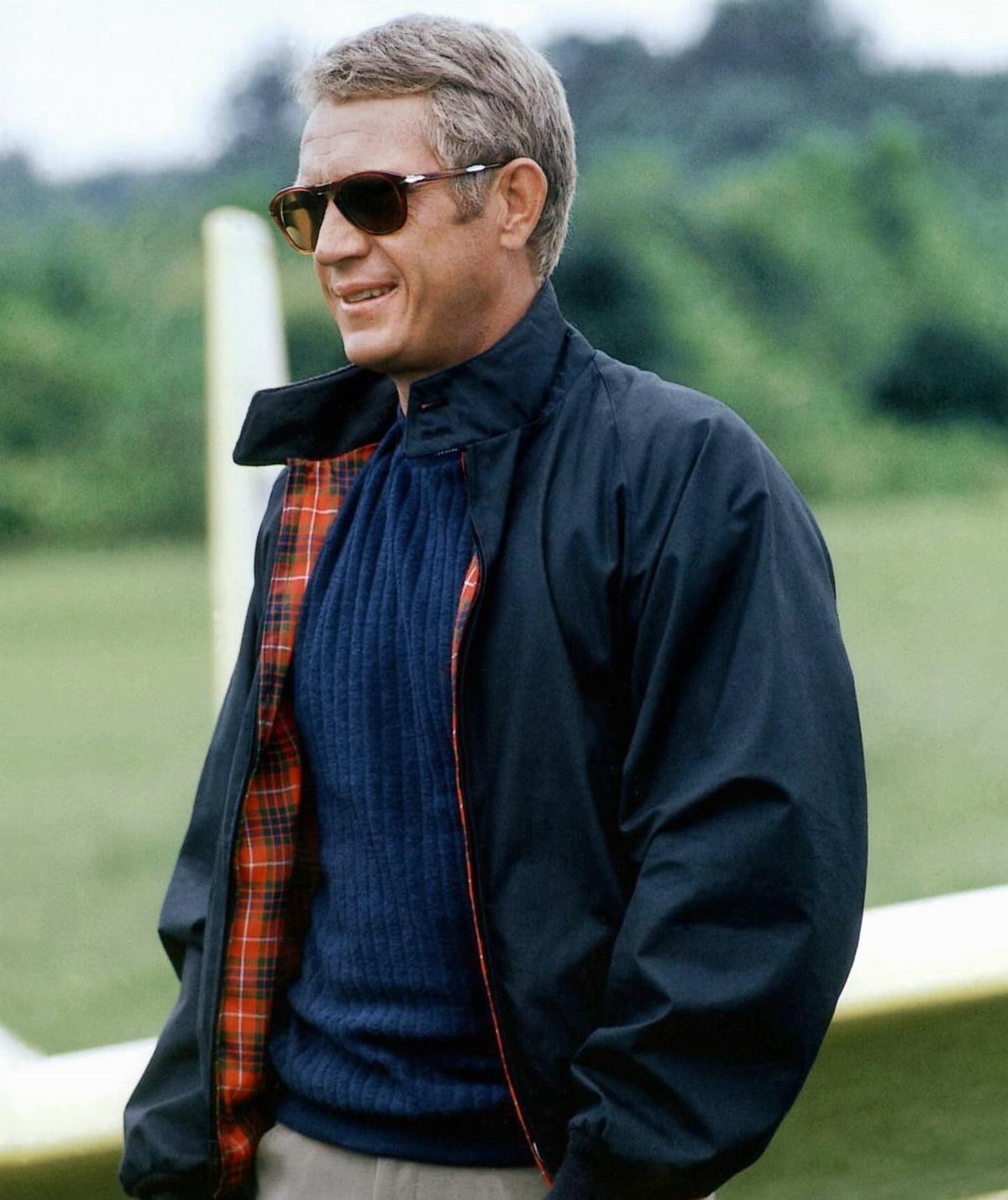 Man in sunglasses and a navy jacket with plaid lining standing outdoors.