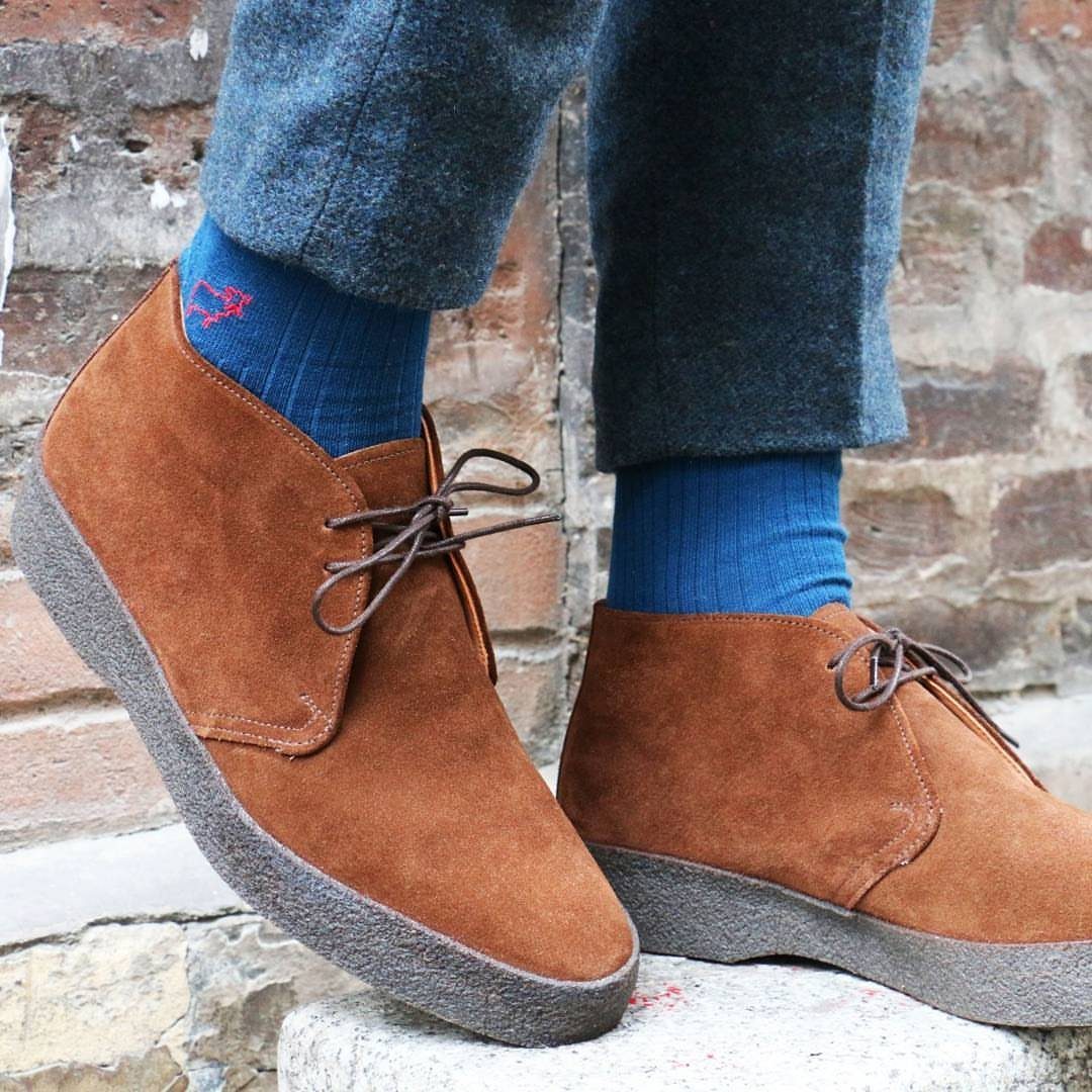 Brown suede chukka boots worn with blue socks and grey trousers on stone steps.