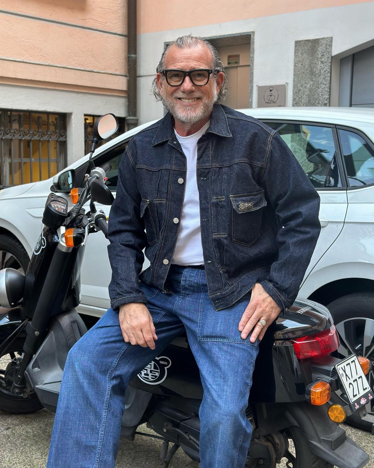 A smiling man with glasses wearing a dark denim jacket and blue jeans sitting on a scooter.