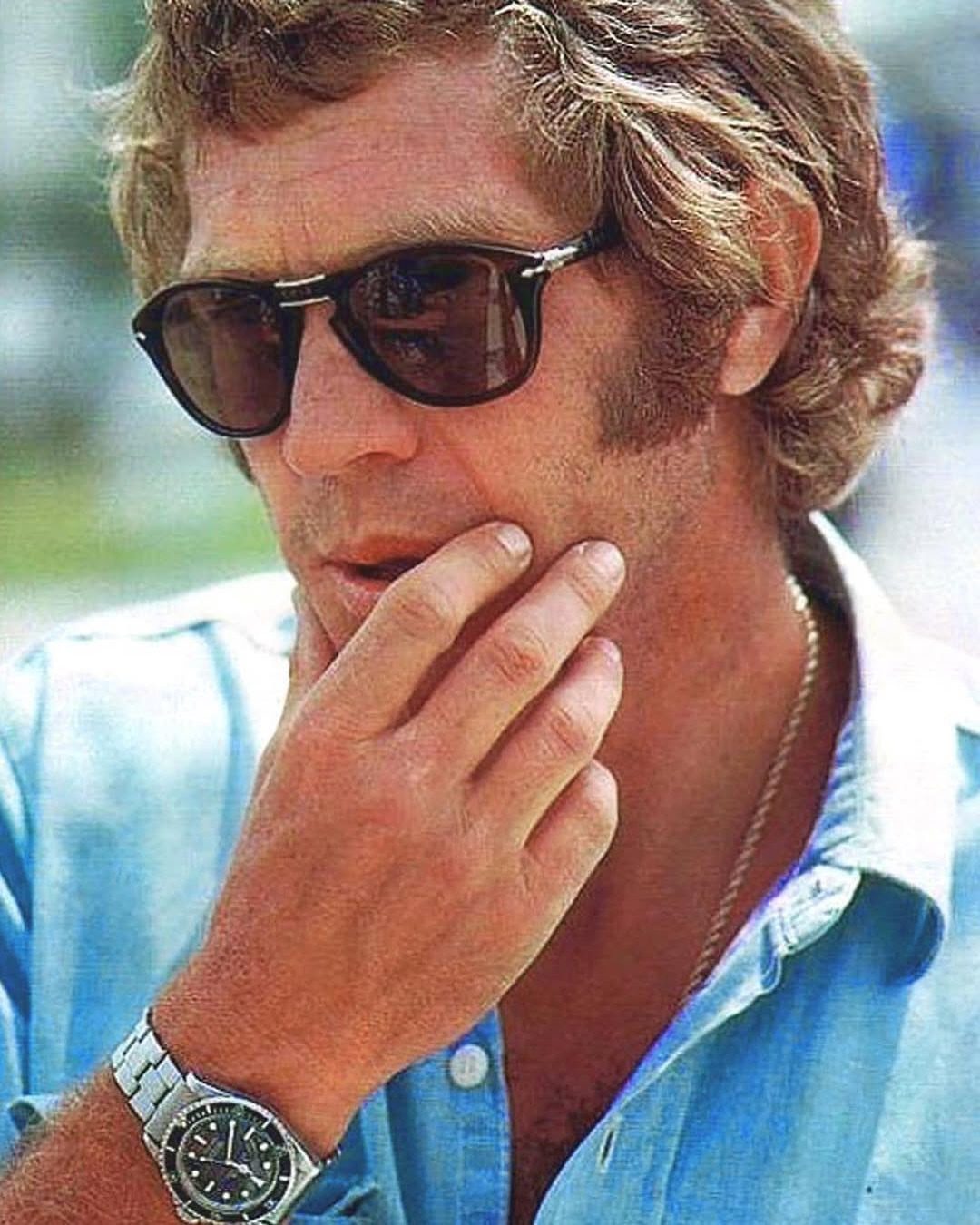 Man with wavy hair wearing dark-tinted sunglasses and a light blue shirt.