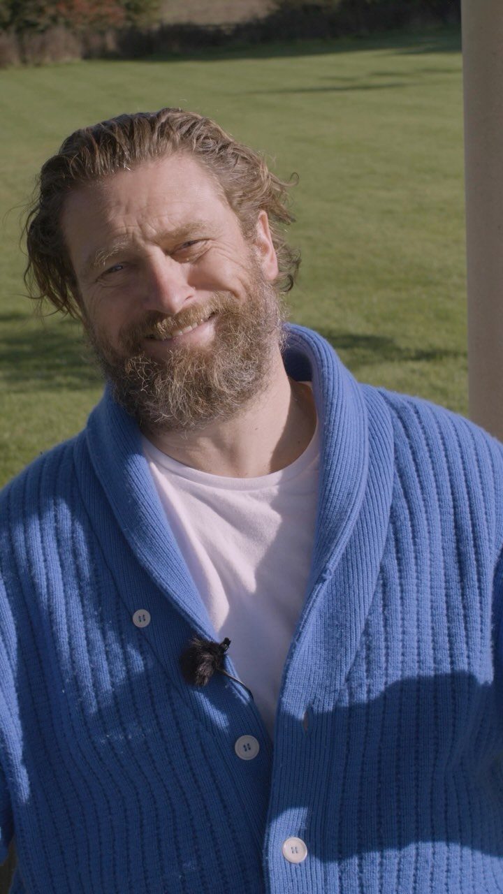 A smiling bearded man wearing a blue ribbed shawl collar cardigan and a pink t-shirt.