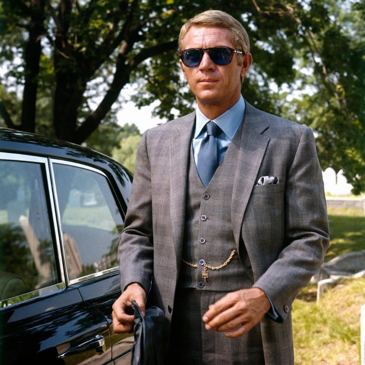 Man in a grey three-piece suit and sunglasses standing next to a black car.