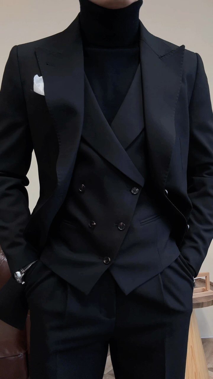 A person wearing a black blazer, waistcoat, and trousers over a black turtleneck.