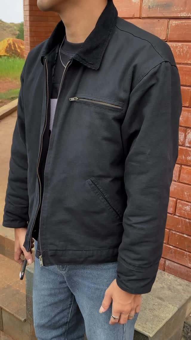 Man wearing a black utility jacket and blue jeans standing by a brick wall.