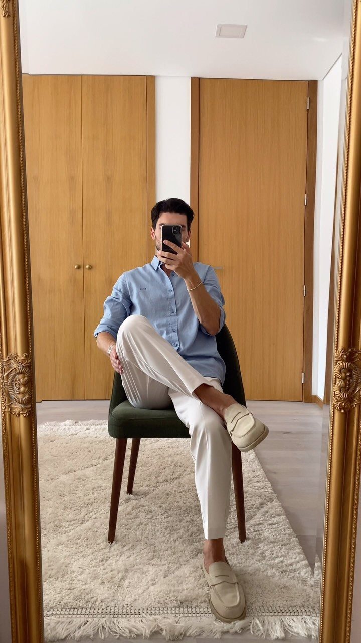 A man in a blue shirt and white trousers wearing cream loafers sitting in a chair.