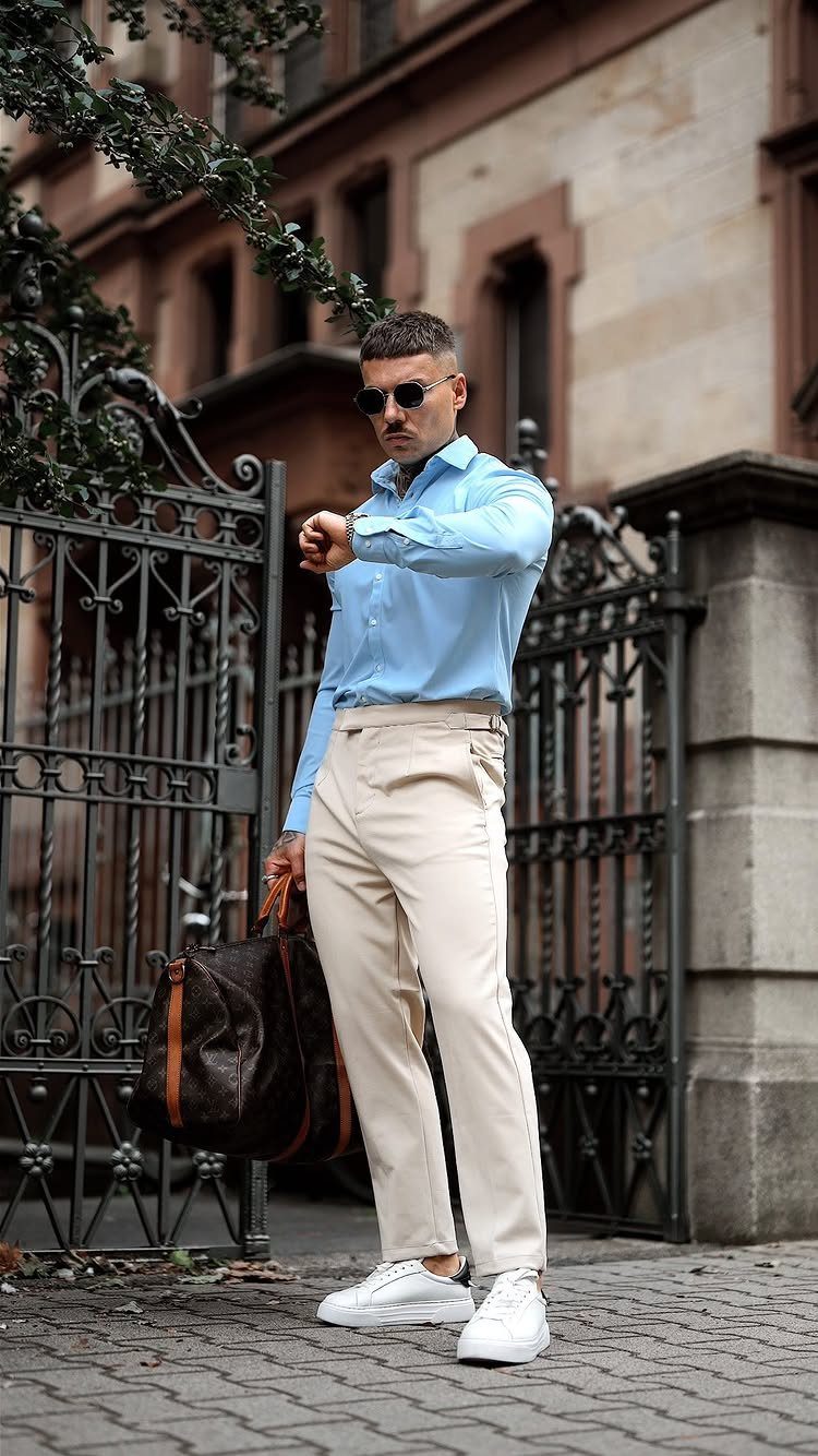 Man in light blue shirt and cream trousers standing by a black metal gate.